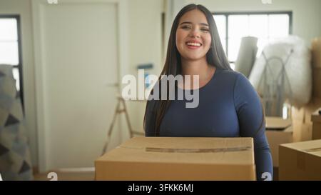 Frau trägt einen Karton in einem Wohnhaus, hält ihn mit beiden Händen und lächelt zwischen gestapelten Umzugskartons und Verpackungsmaterialien; Stockfoto