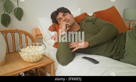 Junger hispanischer Mann lehnt seine Wange auf die Hand, während er in einem Wohnraum in einem Gebäude neben einer Schüssel Popcorn und einer TV-Fernbedienung im Bett liegt; ruhige Langeweile Stockfoto