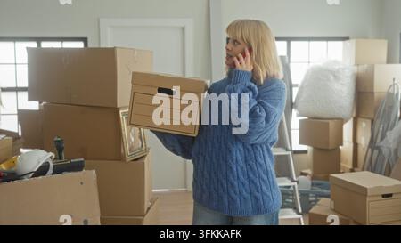 Frau hält einen Karton und spricht am Smartphone, Hand am Ohr, trägt Kartons zwischen gestapelten Verpackungskartons, Leiter und Luftpolsterfolie in einem Gebäude Stockfoto