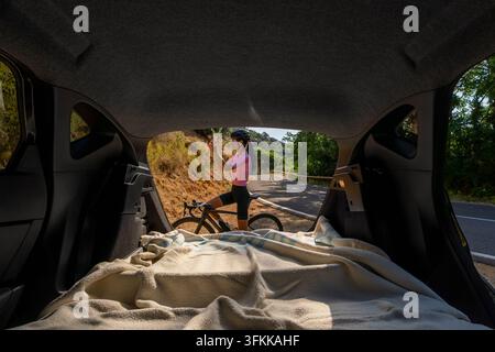 Blick aus dem Kofferraum eines Autos auf eine Radfahrerin auf dem Seitenstreifen der Straße, die eine Radbrille aufsetzt, während sie auf ihrem Rennrad auf der Cost sitzt Stockfoto