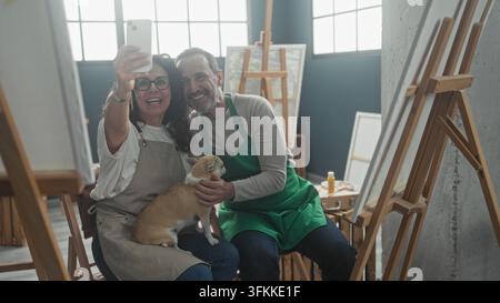Mittlere ältere Frau hält Smartphone und macht ein Selfie mit einem Mann und einem kleinen Hund auf ihrem Schoß im Studio; verspielte Zuneigung. Stockfoto