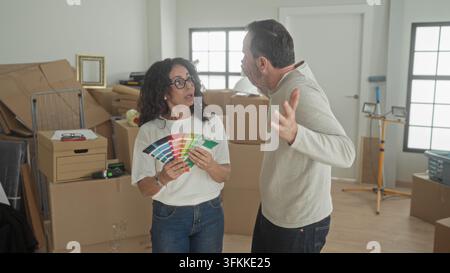 Mittelaltes Paar hält einen Pinselventilator, während die Frau auf einen Farbmuster zeigt, der Mann gestikuliert, umgeben von Kartons und Arbeitsleuchten in einem Gebäude; Haus im Stockfoto