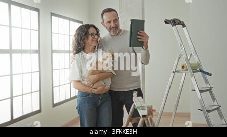 Mann und Frau, mittelaltes Paar, lächeln, während die Frau einen kleinen Hund hält und die Hand des Mannes ein Tablet hält, um ein Selfie in einem Gebäude zu machen; Freude an der Hausrenovierung. Stockfoto
