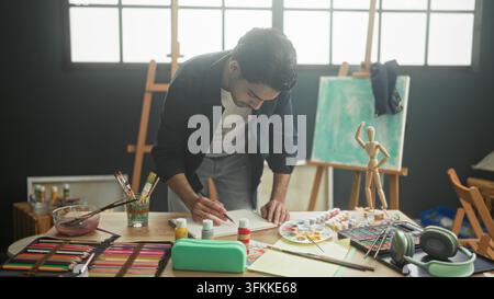 Mann lehnt sich über Tisch und zeichnet mit Bleistift, Hand auf Papier, umgeben von Farben und Holzmannequin im Atelier; kreative Konzentration. Stockfoto