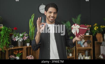 Mann lächelt, während er eine rote herzförmige Geschenkschachtel mit Schleife hält und in einem Blumenstudio das Ok-Zeichen macht; Romantik Wärme. Stockfoto