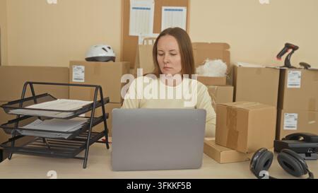 Frau am Schreibtisch sortiert Pakete und Papierkram neben einem Laptop, greift nach einem Dokumentenbehälter und hält die Hand an den Hals in einem Verpackungsgebäude; E-Commerce-Erschöpfung Stockfoto
