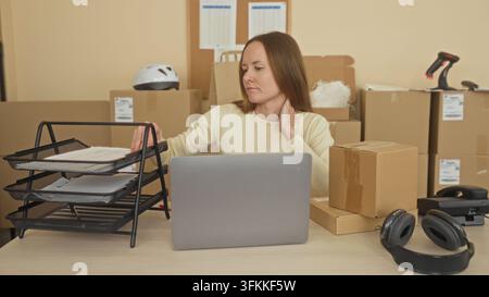 Frau am Schreibtisch sortiert Pakete und Papierkram neben einem Laptop, greift nach einem Dokumentenbehälter und hält die Hand an den Hals in einem Verpackungsgebäude; E-Commerce-Erschöpfung Stockfoto