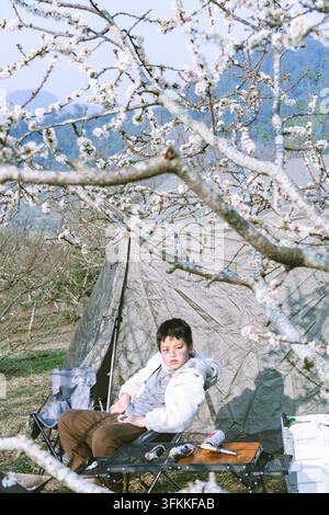 Junge, der in einem Campingstuhl neben einem Zelt unter blühenden Pflaumenbäumen entspannt, Moc Chau, Son La, Vietnam Stockfoto
