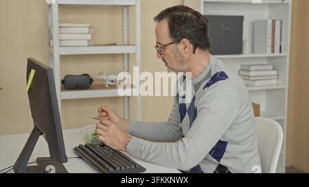 Mann hält Gabel zum Mund, isst Salat am Computer in einem Gebäude, Hände auf Schüssel und Tastatur, trägt Brille und Argyle-Pullover, während er am Schreibtisch sitzt; con Stockfoto