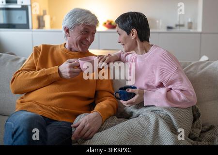 Ein liebevolles Seniorenehepaar trinkt Tee und lächelt sich an, während es zusammen auf dem Sofa zu Hause sitzt Stockfoto