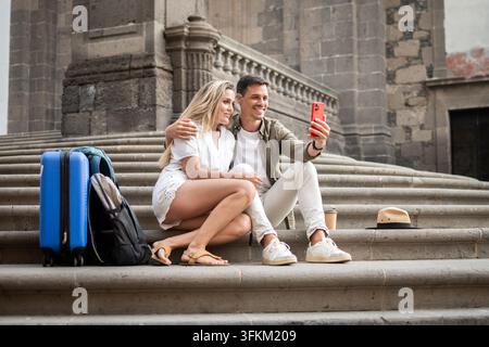 Glückliches junges Paar macht ein Smartphone-Selfie, während es während des Urlaubs mit Koffer und Rucksack auf historischen Stadtstufen sitzt. Stockfoto