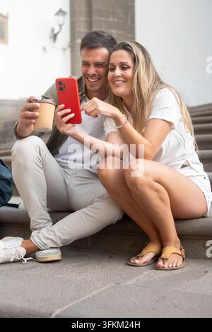 Fröhliches junges Touristenpaar sitzt draußen auf Steinstufen, schaut gemeinsam auf ein Smartphone und genießt einen Kaffee während eines Stadtausflugs. Stockfoto