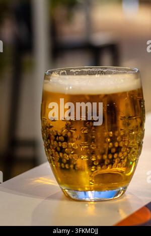 Kaltes Zapfbier, serviert in einem strukturierten Glas mit cremigem Schaumkopf auf einem Bartisch, Nahaufnahme von Getränken in einer gemütlichen Nachtclubsituation. Stockfoto