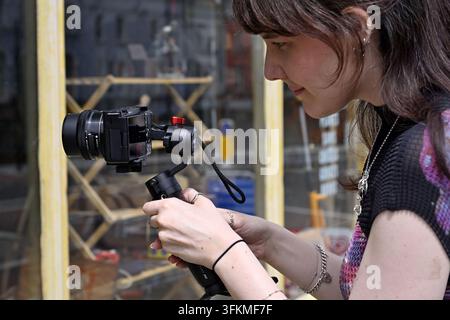Frauenfotografin, Videografin, die eine Handheld-Steadicam benutzt, während sie durch ein Schaufenster filmt, UK Stockfoto