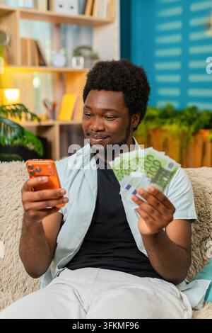Afroamerikanischer junger Mann auf dem Heimsofa zählt Euro-Geld und überprüft sein Smartphone, fühlt sich zufrieden nach einem Online-Spielgewinn. Schwarzer Mann lächelt und überprüft Geld mit dem Mobiltelefon nach einem Sportwettgewinn. Stockfoto