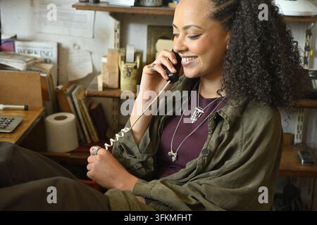 Frau spricht fröhlich am schnurgebundenen Festnetztelefon und sitzt am Arbeitsbereich Stockfoto