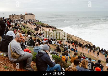 Nazare, Portugal - 6. Dezember 2025: Menschen stehen auf Klippen und beobachten den Ozean und die großen Wellen bei Sonnenuntergang in Nazare, Portugal Stockfoto