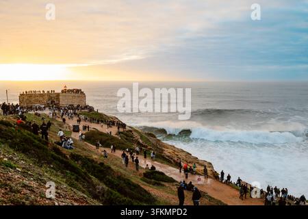 Nazare, Portugal - 6. Dezember 2025: Menschen stehen auf Klippen und beobachten den Ozean bei Sonnenuntergang in Nazare, Portugal Stockfoto