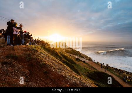 Nazare, Portugal - 6. Dezember 2025: Menschen stehen auf Klippen und beobachten den Ozean und die großen Wellen bei Sonnenuntergang in Nazare, Portugal Stockfoto