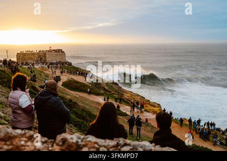 Nazare, Portugal - 6. Dezember 2025: Menschen stehen auf Klippen und beobachten den Ozean und die großen Wellen bei Sonnenuntergang in Nazare, Portugal Stockfoto