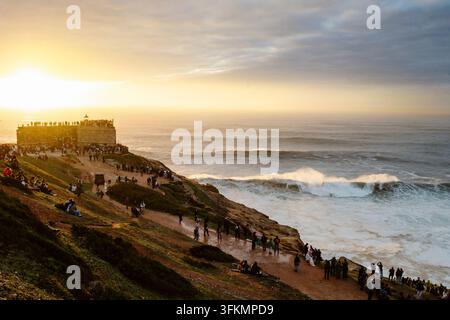 Nazare, Portugal - 6. Dezember 2025: Menschen stehen auf Klippen und beobachten den Ozean und die großen Wellen bei Sonnenuntergang in Nazare, Portugal Stockfoto