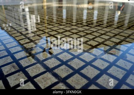 Nahaufnahme einer mit flachem Wasser überfluteten Granit-Mosaikflächte, die spiegelähnliche Reflexionen der umgebenden städtischen Umgebung erzeugt Stockfoto