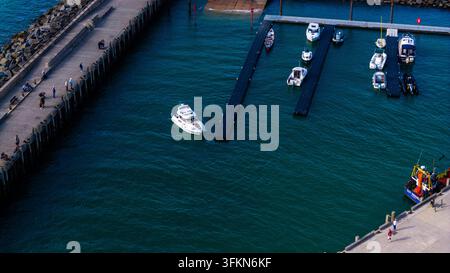 Luftaufnahme des West Bay Hafens mit verschiedenen Booten, die an schwimmenden Stegen festgemacht sind, einem kleinen Motorboot, das durch das ruhige blaue Wasser navigiert, und Menschen, die entlanggehen Stockfoto