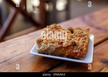 Nahaufnahme eines Stücks gebackener Pfirsich- oder Pflaumen-Crumble-Kuchen, serviert auf einem kleinen quadratischen weißen Teller, der auf einem Holztisch steht. Stockfoto
