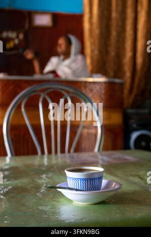 Eine Tasse Kaffee ruht auf einer Untertasse mit einem Löffel auf einem grünen Tisch vor einem leeren Metallstuhl, während eine verschwommene Frau, die in einen weißen Schal gewickelt ist, sitzt b Stockfoto