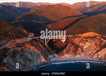 Luftaufnahme zeigt die Bixby-Creek-Brücke entlang des Pacific Coast Highway in Big Sur, Kalifornien, die einen felsigen Küstencanyon unter warmem Sonnenlicht überspannt Stockfoto