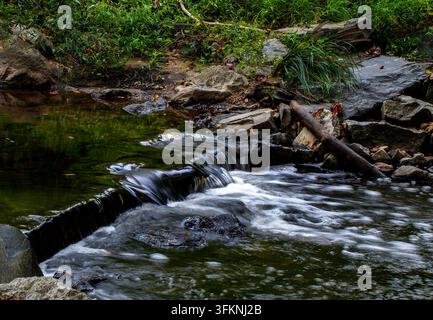 Ein kleiner, flacher Bach oder Fluss, der über eine Reihe von niedrigen, stufenartigen Steinabfällen oder kleinen Felsbändern fließt, umgeben von grüner Vegetation und Felsen Stockfoto