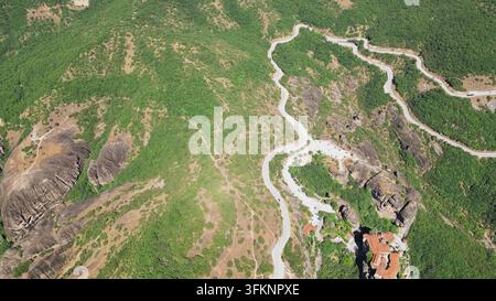 Horizontale Luftaufnahme einer sich windenden Bergserpentinstraße und eines Klostergebäudes zwischen einzigartigen Felsformationen und grünen Hügeln in Meteora, Griechenland. Stockfoto