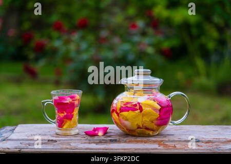 Schöner und duftender Tee aus gelben, rosa und roten Rosenblüten in einer Glas-Teekanne und einer Tasse im Sommergarten auf einem Holztisch. Nahaufnahme von Kräuterblüten Stockfoto