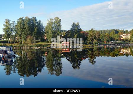 Ein ruhiger See mit Bäumen und einem kleinen roten Gebäude am Ufer, das seine Spiegelungen im Wasser zeigt, Bengtsfors, Dalsland, Dalslandkanal, Schweden Stockfoto