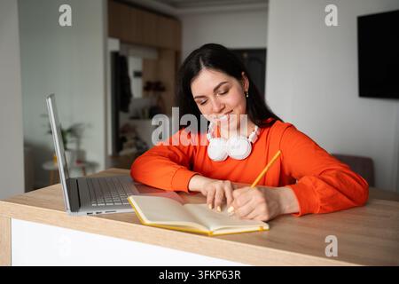 Junge Frau macht Notizen neben ihrem Laptop zu Hause, während sie an einem Online-Kurs teilnimmt. Fernstudium, Bildung, Produktivität und Selbstorganisation. Stockfoto
