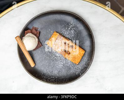 Ein Apfelstrudel, serviert mit einer Kugel Vanilleeis, einem zigarrenförmigen Keks und Schokoladenraspeln auf einem schwarzen Teller in Tienen. Die Dessert-Kombi Stockfoto