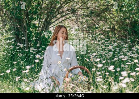 Eine Frau in einem weißen Gewand sitzt auf einer Waldlichtung in einem Feld voller Gänseblümchen und hält einen Holzkorb. Stockfoto