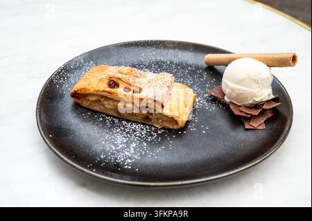 Ein Apfelstrudel, serviert mit einer Kugel Vanilleeis, einem zigarrenförmigen Keks und Schokoladenraspeln auf einem schwarzen Teller in Tienen. Das Dessert kombiniert warmes Gebäck mit Eiscreme und dekorativen Schokoladenelementen. Tienen, Belgien 19. SEP 2026 Foto Werner Lerooy. Stockfoto