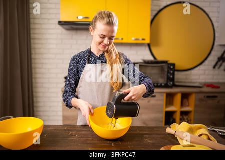 Junge Frau gießt Teig in eine Rührschüssel in einer warmen Küche mit gelben Akzenten Stockfoto