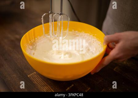 Hände schlagen Eiweiß in einer gelben Rührschüssel auf einem Holztisch unter warmem Küchenlicht Stockfoto