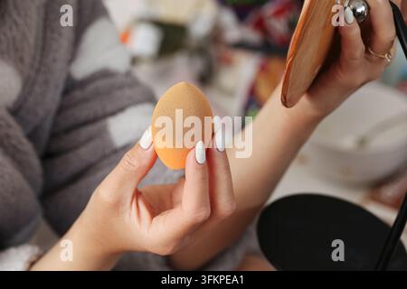 Frau hält orangefarbenen Make-up-Schwamm neben Kompaktspiegel. Schönheitshintergrund für die Anwendung von Kosmetikgrundierung zum Mischen von Gesichtspflege und Make-up-Routine Stockfoto