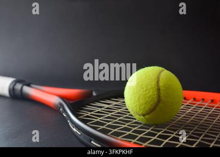 Ein gelber Tennisball ruht auf den Saiten eines roten und schwarzen Schlägers, mit einem weiteren Schläger dahinter vor einem dunklen Studiohintergrund. Stockfoto