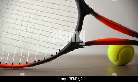 Ein roter und schwarzer Tennisschläger dominiert den Vordergrund, während ein gelber Ball im Hintergrund sitzt und Tiefe, Perspektive und diagonale Komposition betont Stockfoto