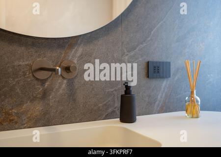 Zeitgenössisches italienisches Design in einem angeschlossenen Badezimmer mit rundem Spiegel, wandmontiertem Wasserhahn, weißem integrierten Waschbecken, Seifenspender und Raumduft-Diffuser. Stockfoto
