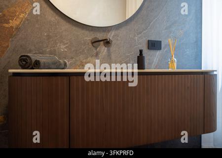 Modernes italienisches Badezimmer mit angrenzendem WC, gerippter freistehender Holzwaschtisch, großer runder Spiegel, dunkler wandmontierter Wasserhahn, Handtücher und Schilfduftspender. Stockfoto