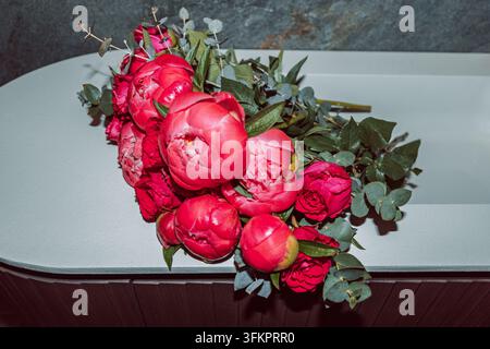 Rosa Pfingstrosen und rote Rosen füllen ein weißes Waschbecken in einem italienisch gestalteten Badezimmer, mit Eukalyptusblättern, einer dunklen Wand und scharfen Blitzschatten. Stockfoto