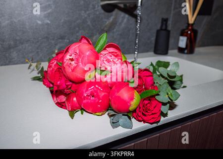 Rosa Pfingstrosen und rote Rosen füllen ein weißes Waschbecken, während Wasser aus einem schwarzen Wasserhahn dahinter in einem zeitgenössisch italienisch gestalteten Badezimmer fließt. Stockfoto