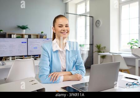 Businessfrau-Porträt mit Laptop im Büro. Junge Berufstätige sitzt an einem aufgeräumten Arbeitsplatz und überprüft eine Finanzanalyse auf einem Computer mit Diagrammen auf s Stockfoto