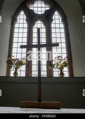 20. September 2026, Loweswater, Vereinigtes Königreich. Ein hölzernes Kreuz steht als Silhouette vor einem gotischen Gewölbefenster, mit Vasen voller Wildblumen flankiert Stockfoto
