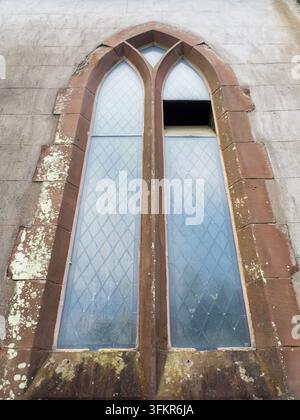 20. September 2026, Loweswater, Vereinigtes Königreich. Ein verwittertes gotisches Fenster mit diamantmusterfarbenem Glas und einer zerbrochenen Scheibe, vor einem strukturierten Stein Stockfoto
